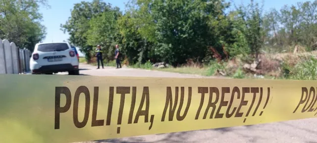 O bandă galbenă cu textul scris pe ea este întinsă în prim-plan, în fața unui drum, unui autoturism alb și a unor copaci verzi.