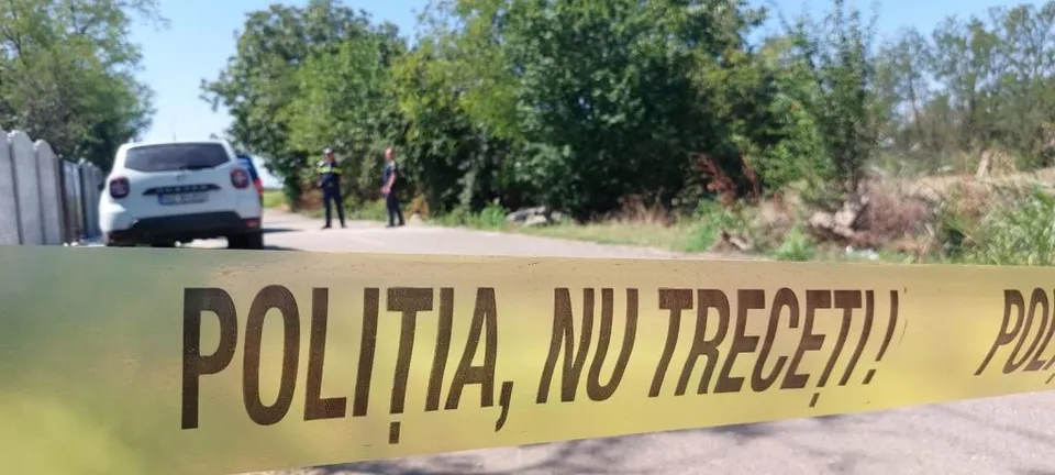O bandă galbenă cu textul scris pe ea este întinsă în prim-plan, în fața unui drum, unui autoturism alb și a unor copaci verzi.