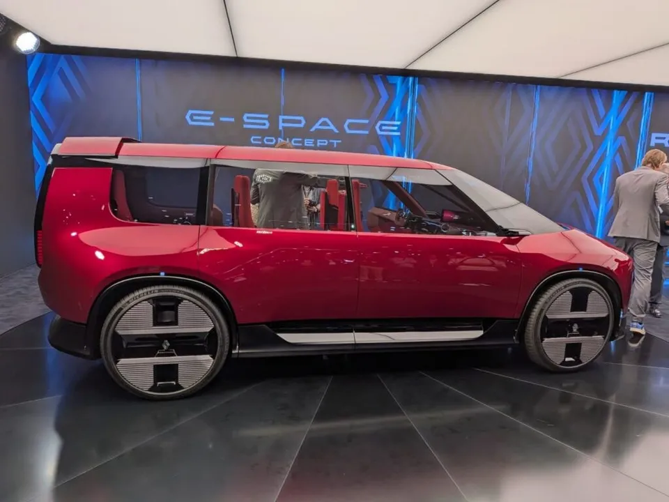 Un concept de vehicul roșu cu ferestre mari este expus într-un spațiu de expoziție cu un panou de fundal albastru pe care scrie text alb.