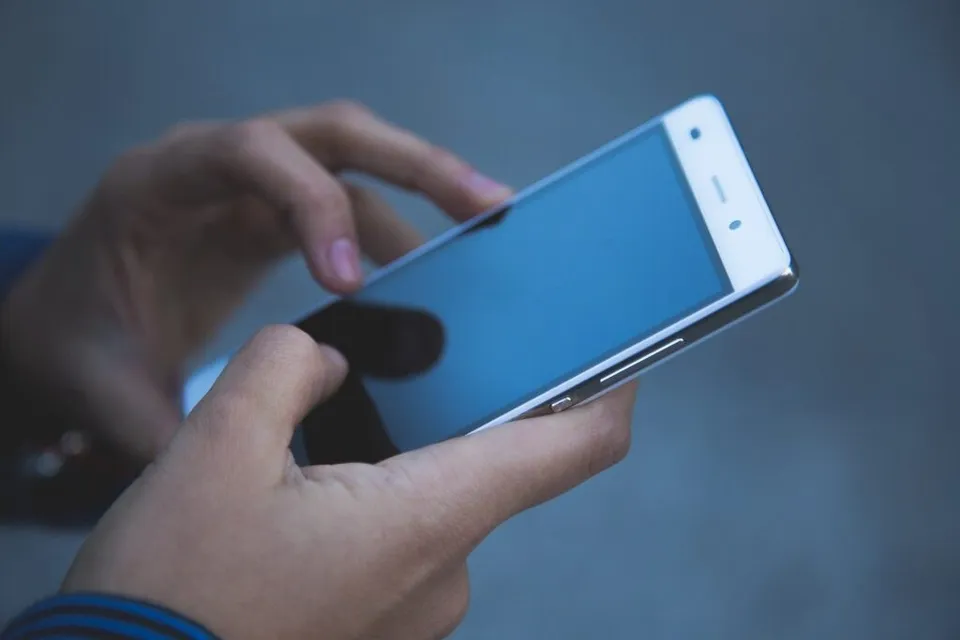 Două mâini țin și utilizează un smartphone cu ecranul albastru pe un fundal gri închis.