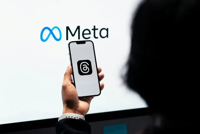 O persoană ține un telefon mobil în față, afișând un logo negru pe ecran, în fața unui ecran mare cu logoul Meta pe fundal alb.