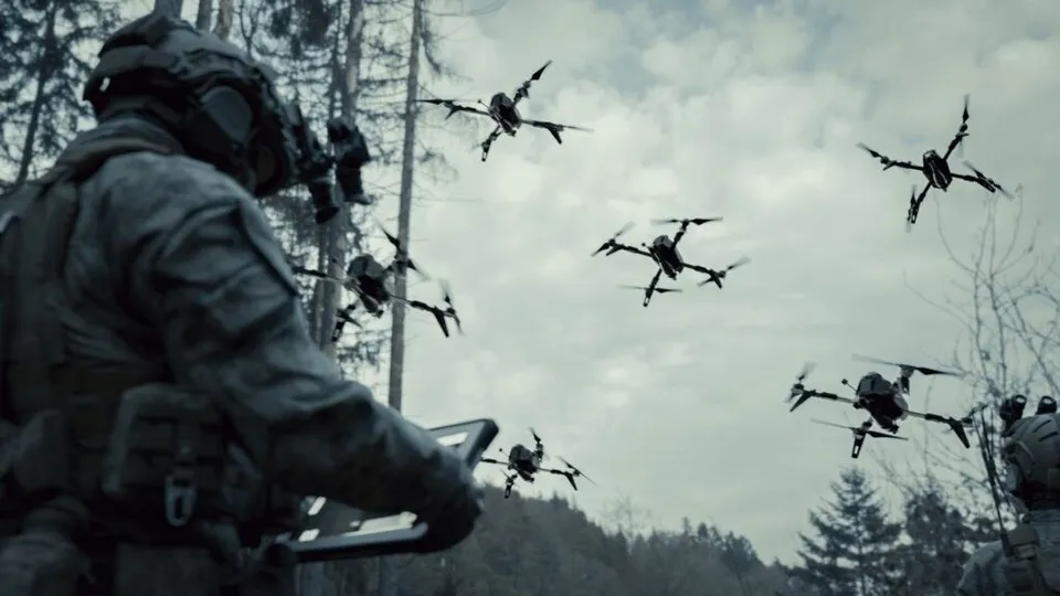 Un soldat în uniformă ține un dispozitiv electronic, privind spre mai multe drone care zboară pe cerul înconjurat de copaci.