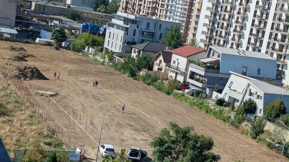 O fetă cu tricou alb merge pe un teren gol și uscat, lângă o clôtură, având clădiri și mașini în fundal.