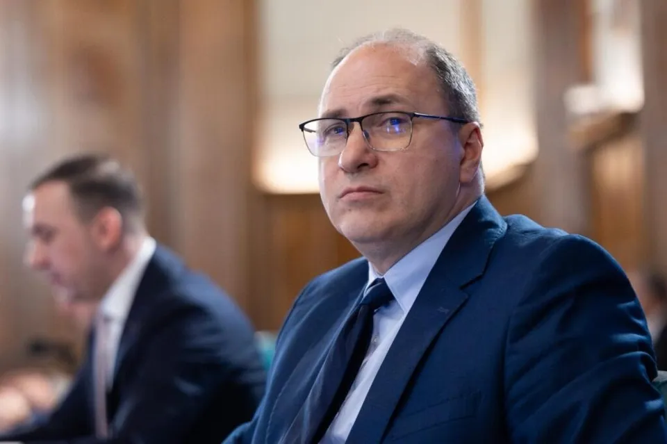 Un bărbat cu ochelari și costum în bleu portocaliu stă într-o încăpere cu fundal de lemn, în timp ce un alt bărbat este vizibil în plan secund.