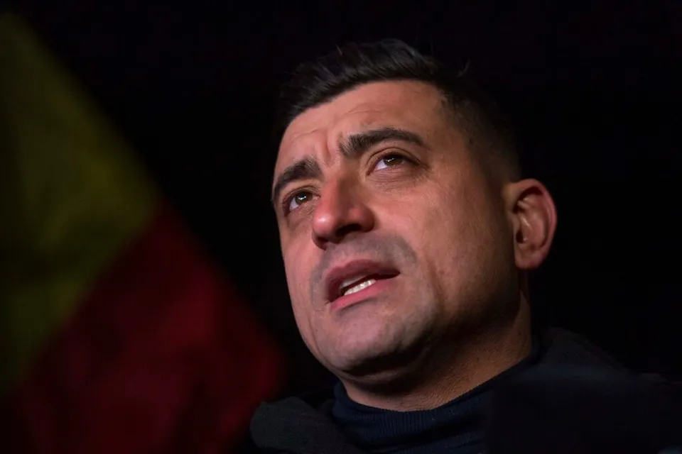 Un bărbat cu părul scurt și haine închise la culoare privește în sus pe un fundal întunecat cu un steag colorat în stânga.