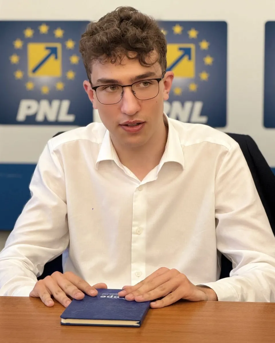 Un bărbat cu părul creț și ochelari, purtând o cămașă albă, stă la o masă cu un obiect dreptunghiular albastru în față.