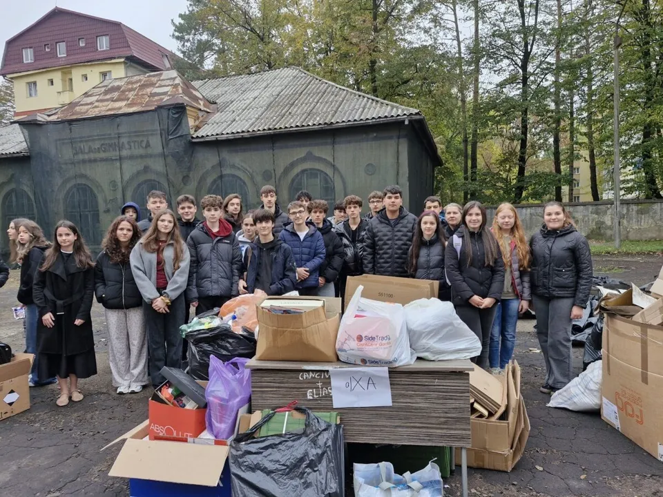 Un grup de elevi în picioare în spatele unei cantități mari de cutii de carton, saci de gunoi și deșeuri reciclabile colectate.