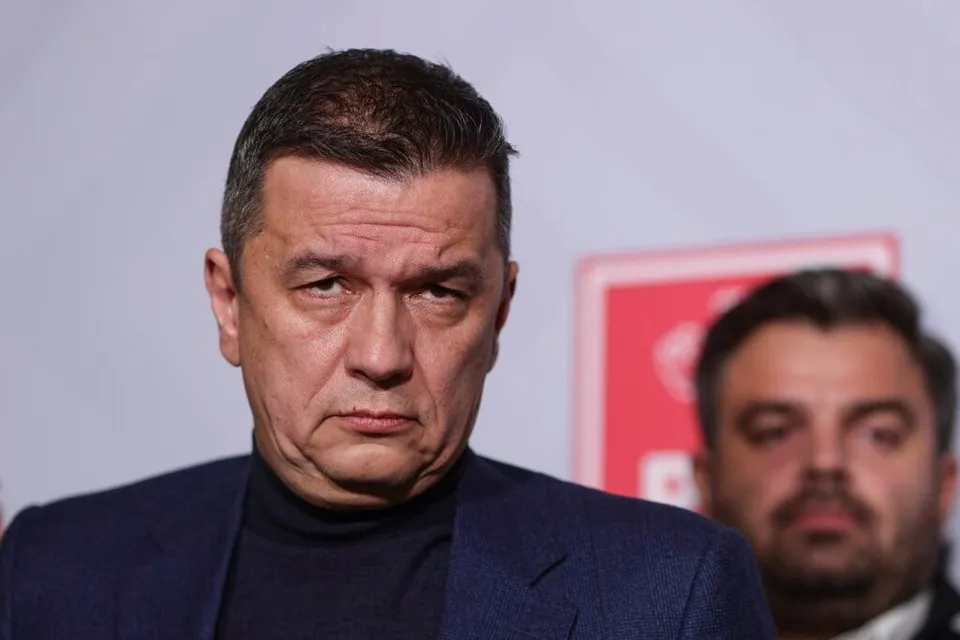 Un bărbat cu părul scurt și întunecat poartă un sacou albastru peste un tricou cu guler negru, privind spre camera foto.