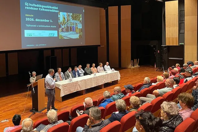 Oamenii stau așezați în spectatori în fața unei mese lungi pe care se află mai mulți bărbați într-o sală de conferințe cu un ecran proiectat.