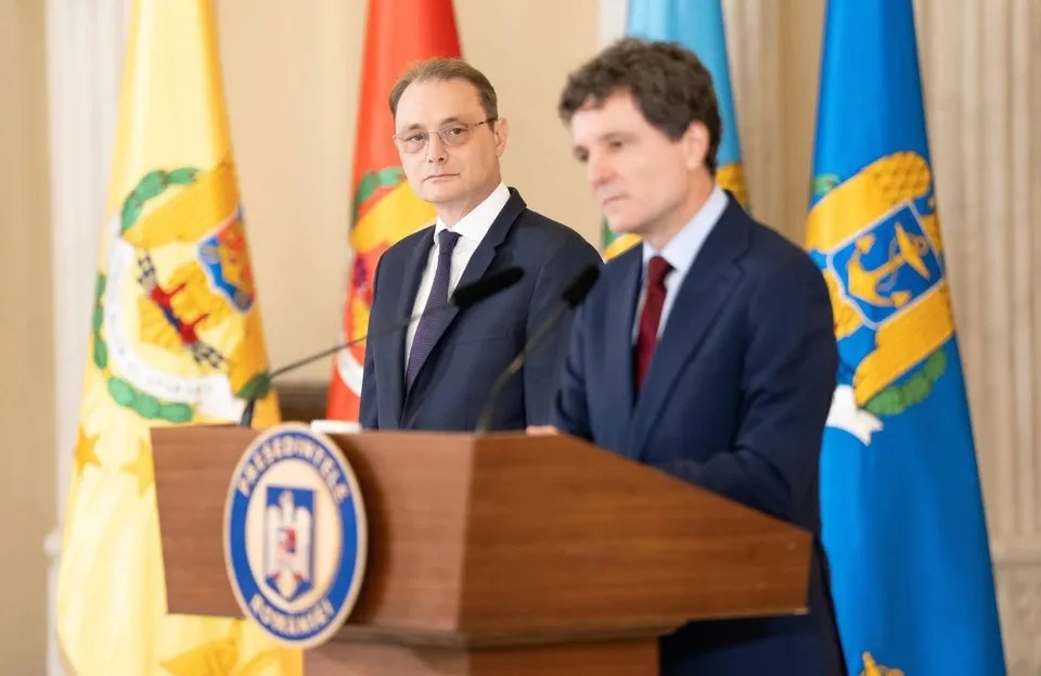 Doi bărbați în costume și cravate stau în spatele unui podium de lemn în fața unor steaguri colorate.