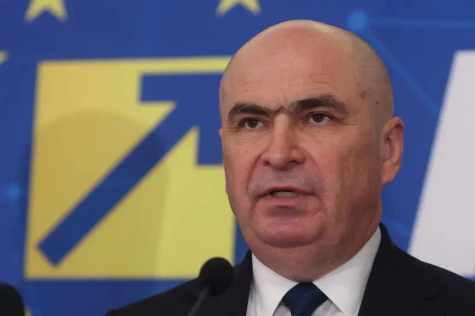 Un bărbat cu părul puțin, purtând cămașă albă, cravată albastră și sacou negru, este surprins în prim-plan în fața unui fundal albastru cu elemente galbene.