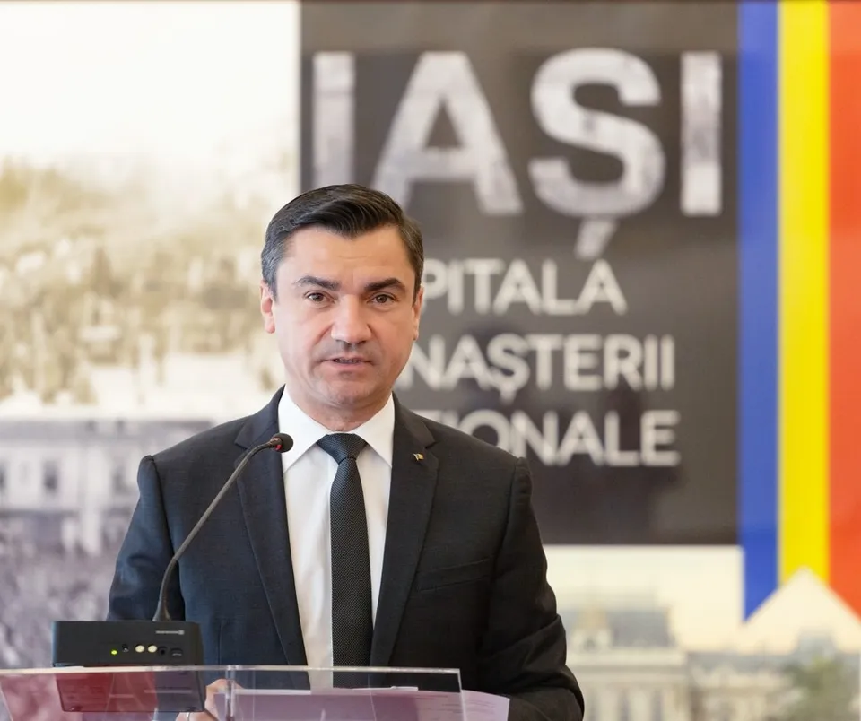 Un bărbat cu părul scurt, purtând sacou negru, cămașă albă și cravată neagră, stă în fața unui microfon în fața unui panou cu textul IAȘI.