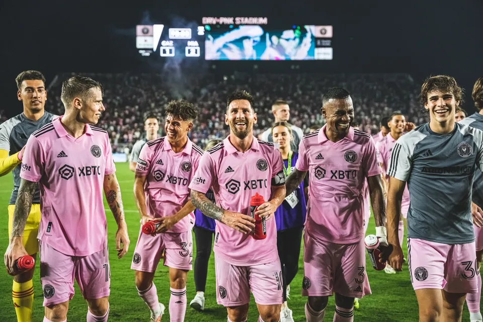 Câțiva jucători de fotbal în echipamente roz merg pe terenul de sport sub luminile stadionului.