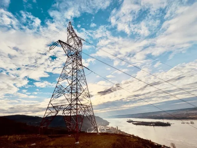 O turn de înaltă tensiune din metal se ridică pe un deal sub un cer cu nori deasupra unui râu și unor dealuri întunecate.