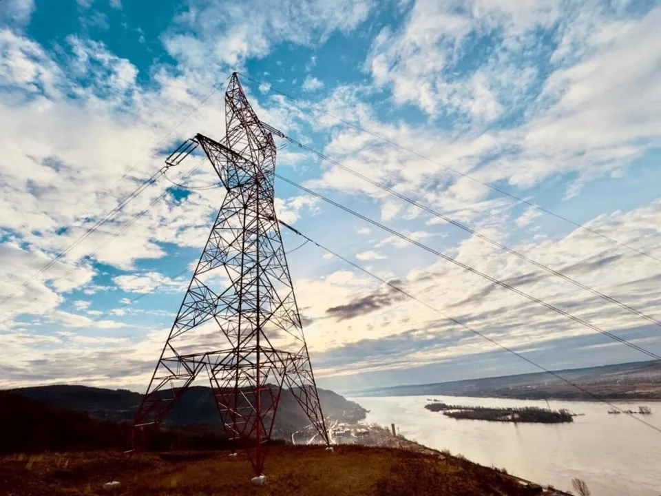 O turn de înaltă tensiune din metal se ridică pe un deal sub un cer cu nori deasupra unui râu și unor dealuri întunecate.