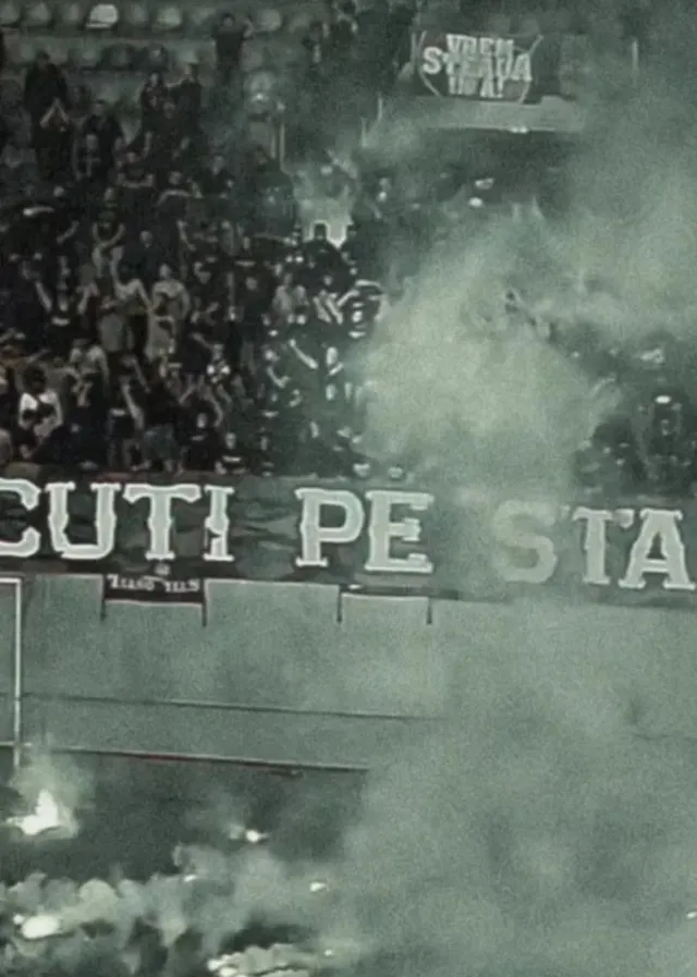 O mulțime de oameni stă pe tribunele unui stadion, acoperite de fum dens și de un banner alb cu litere negre.