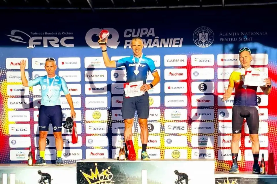 Trei bărbați în echipamente de ciclism stau pe un podium în fața unui panou cu diverse logo-uri de sponsori.