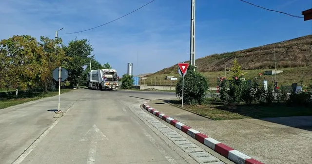 Un camion de gunoi circulă pe un drum asfaltat lângă un deal de pământ sub un cer senin.