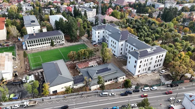 O vedere aeriană prezintă mai multe clădiri albe cu acoperișuri gri și un teren de fotbal verde înconjurat de copaci și drumuri cu mașini.