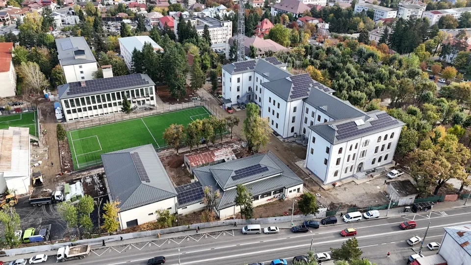 O vedere aeriană prezintă mai multe clădiri albe cu acoperișuri gri și un teren de fotbal verde înconjurat de copaci și drumuri cu mașini.