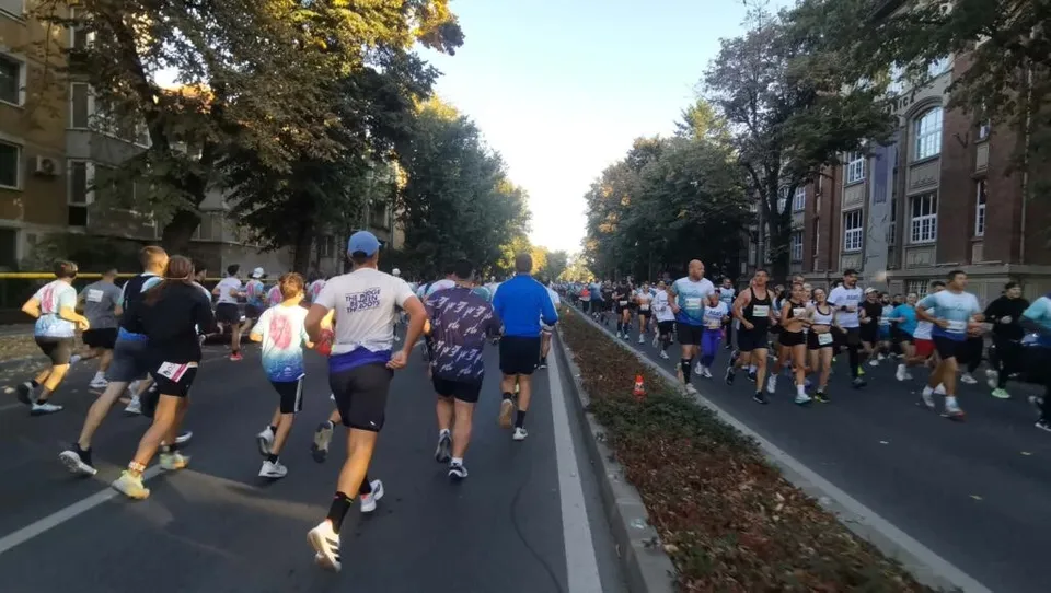 Mulți oameni în haine sportive aleargă pe un drum asfaltat flancat de copaci și clădiri de un etaj sau mai mult.