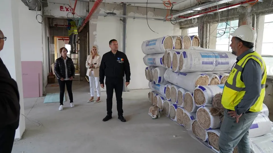 Patru persoane stau într-un spațiu interior cu pereți albi, lângă stive de materiale de izolație învelite în folie albă și albastră.