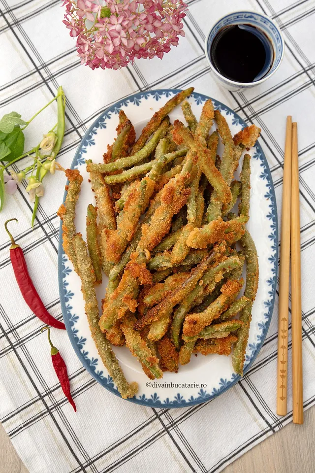 O farfurie albastră și albă plină cu fasole verde prăjită, crocantă, alături de două ardei roșii, niște bețe de chopstick, o floare roz și o cană cu sos negru.