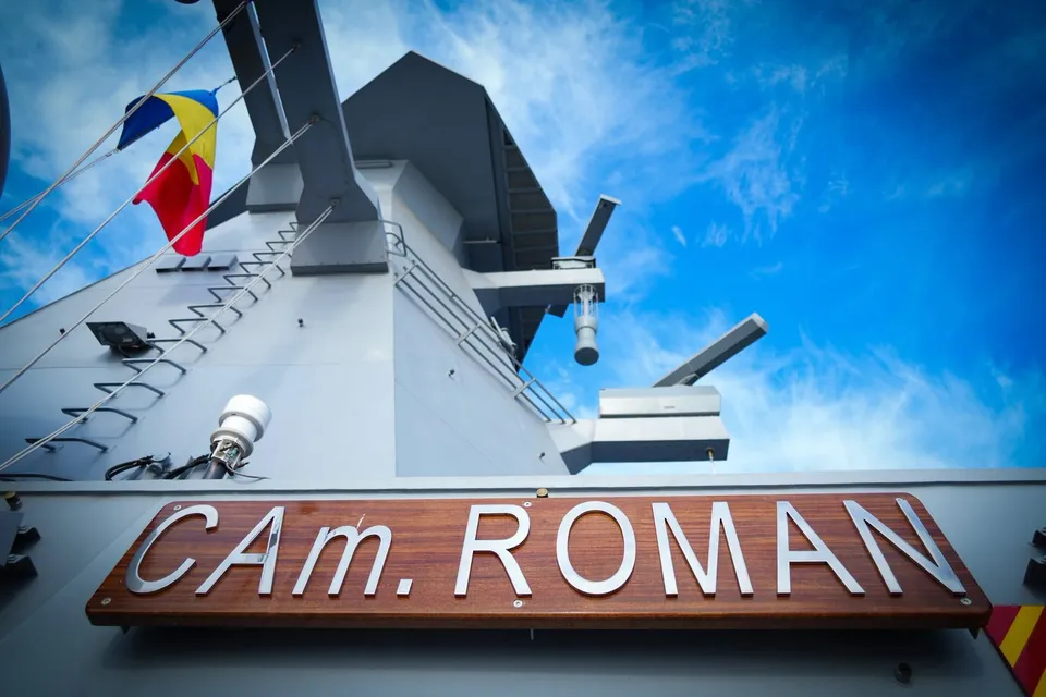 O placă de lemn cu textul metalic "CA. ROMAN" este montată pe suprafața unei nave gri sub un cer albastru.