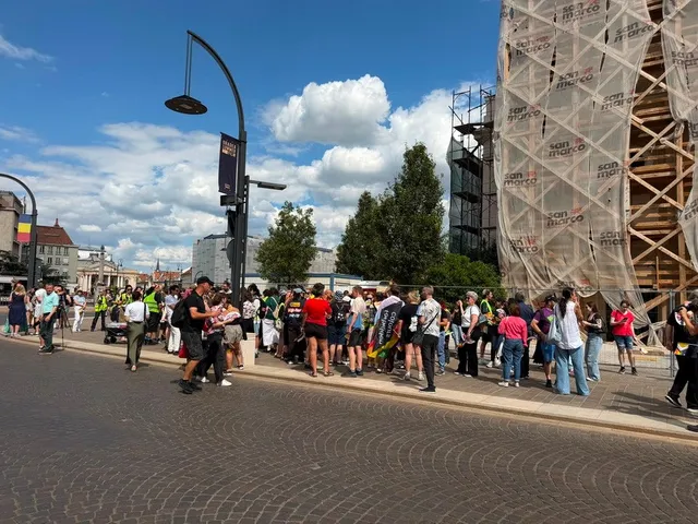 Un grup de persoane stă adunat pe un trotuar lângă o clădire acoperită de un panou de șantier sub un cer cu nori albi.