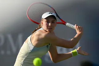 O femeie cu șapcă albă și echipament sportiv de culoare deschisă lovește o minge de tenis cu o rachetă într-un teren de tenis.