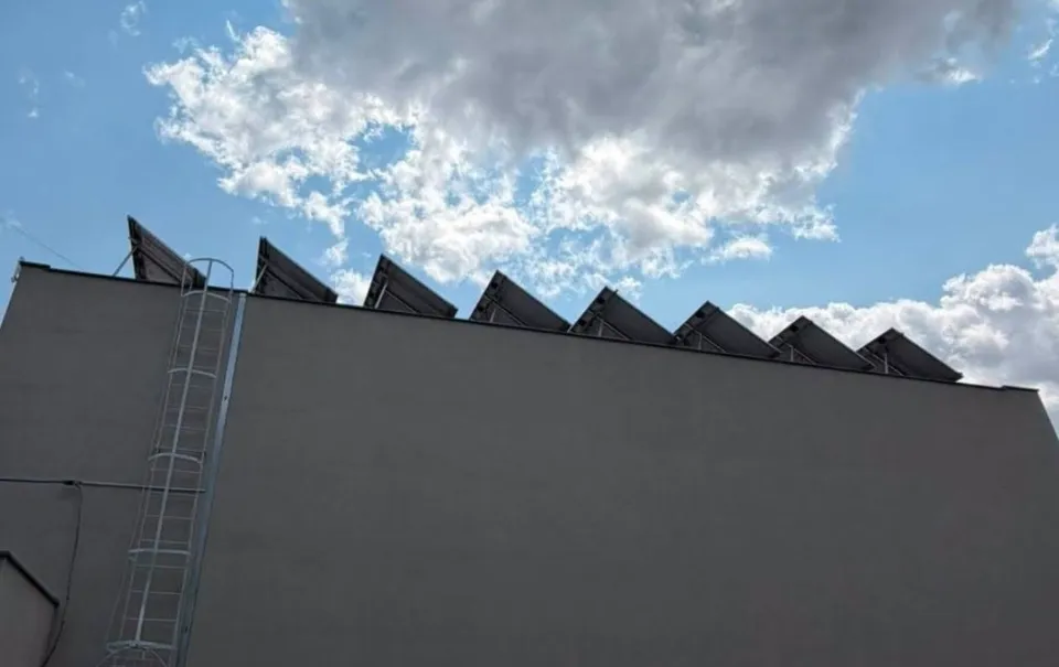 Mai multe panouri solare sunt instalate pe marginea unei acoperișuri gri sub un cer cu nori albi și albastre.