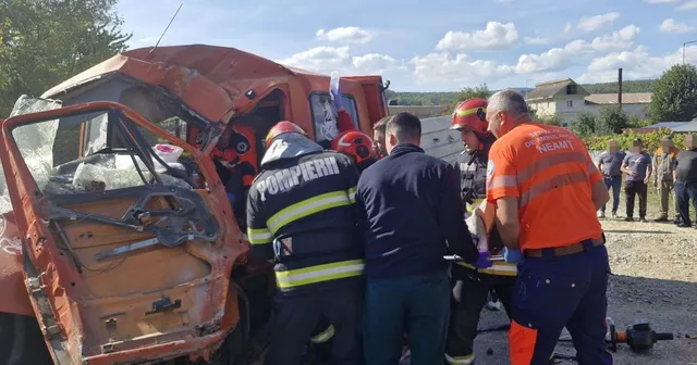 Mai mulți salvatori în uniforme de lucru și casci de protecție lucrează lângă o autoutilitară de culoare portocalie puternic avariată.