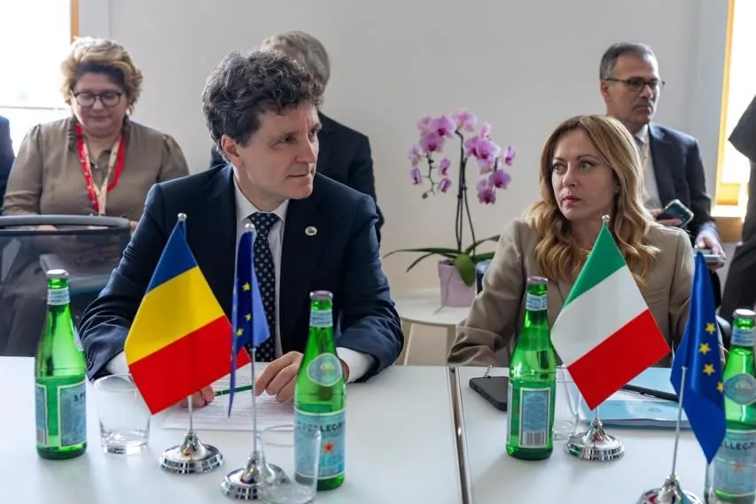Un bărbat într-un costum închis și o femeie într-o jachetă deschisă stau la o masă cu steaguri mici ale României, Italiei și Uniunii Europene.