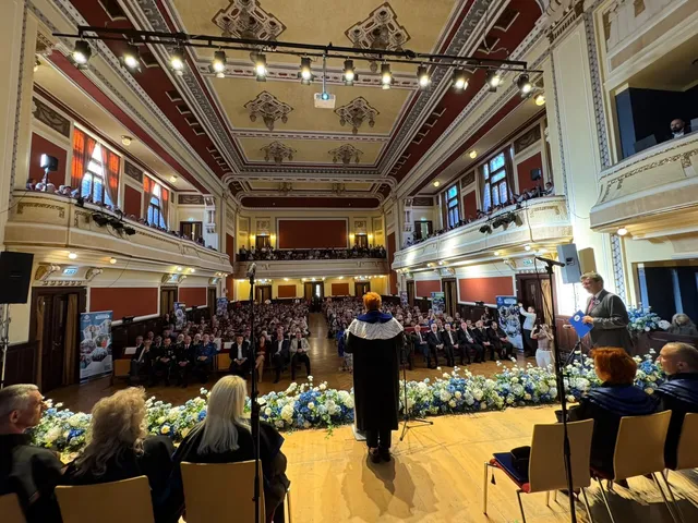 Oamenii stau așezați în scaune în fața unei tribune de lemn, într-o sală de ceremonie cu tavan înalt și decoruri ornamentale.