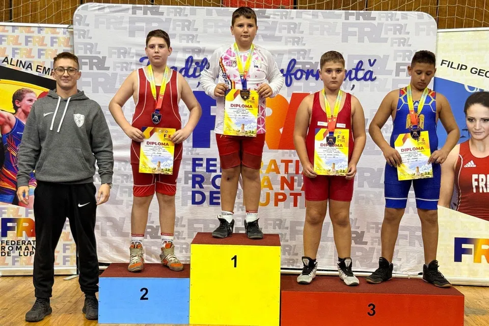 Cinci băieți stau pe un podium colorat, patru dintre ei purtând medalii și diplome, în timp ce un bărbat stă în partea stângă.