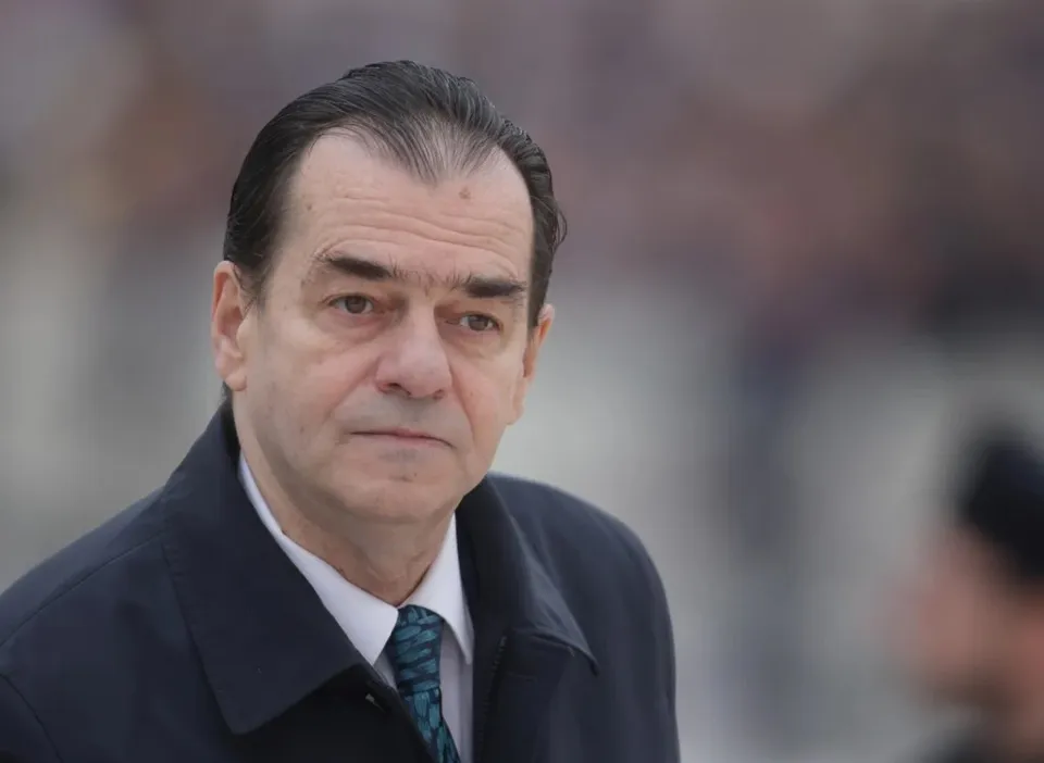 Un bărbat cu părul scurt și închis la culoare poartă o jachetă neagră, cămașă albă și cravată cu modele verzi.