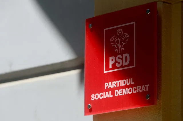 Un panou roșu dreptunghiular cu textul alb și un simbol de trei flori este montat pe un perete galben.