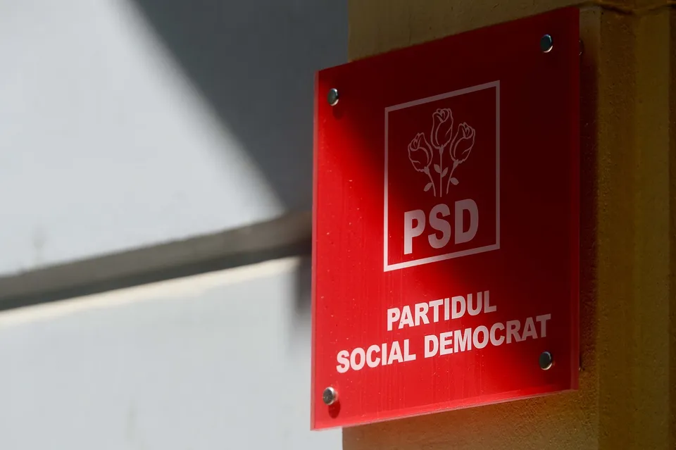 Un panou roșu dreptunghiular cu textul alb și un simbol de trei flori este montat pe un perete galben.