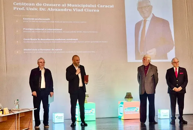 Patru bărbați stau pe un podium în fața unui ecran mare care afișează un portret și un text.
