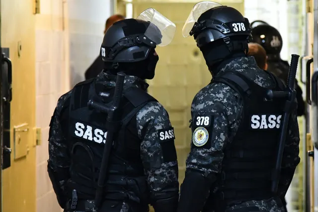 Trei persoane purtând uniforme de culoare gri cu modele de camuflaj și echipament tactice negru stau într-un coridor cu pereți deschiși.