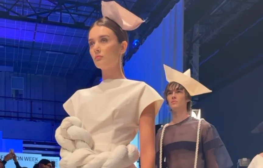 Două persoane poartă haine albe și gri cu accesorii geometrice pe cap și elemente de sfoară într-un spațiu iluminat cu lumini albastre.
