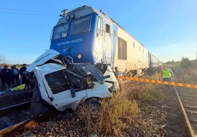 O locomotivă albastră și gri este oprită lângă resturile unei autoutilitare albe zdrobite pe o cale ferată, în timp ce persoane stau în apropiere.