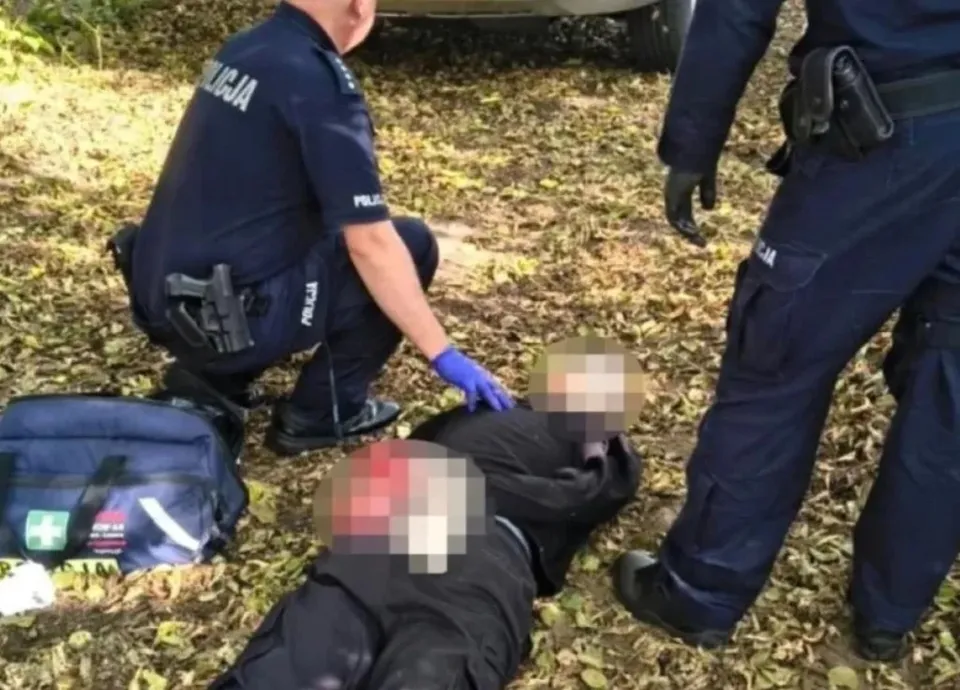 Un polițist cu mănuși albastre stă ghemuit lângă două persoane întinse pe iarbă, lângă o geantă medicală albastră.