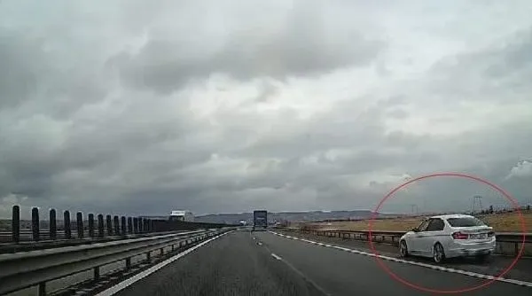 O mașină albă circulă pe o autostradă sub un cer înnorat, lângă o barieră metalică de protecție.