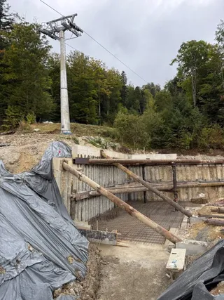 Un pilon metalic de telegondolă se ridică lângă un șantier cu suporturi de lemn și materiale acoperite cu folie gri sub un cer înnorat.