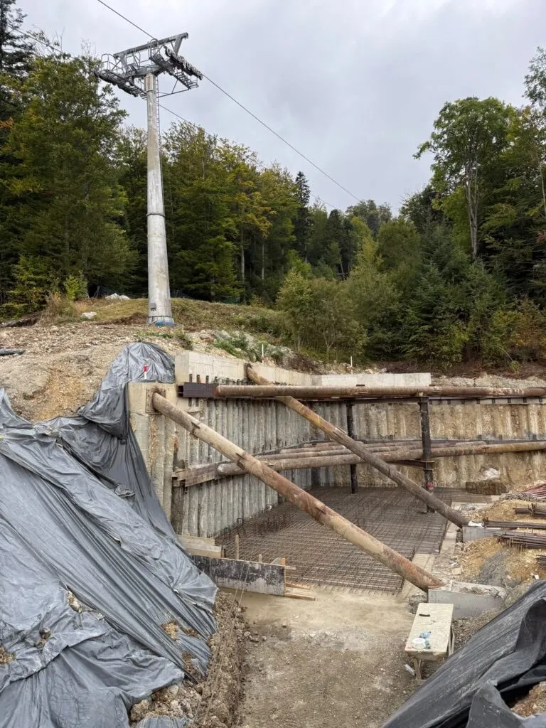 Un pilon metalic de telegondolă se ridică lângă un șantier cu suporturi de lemn și materiale acoperite cu folie gri sub un cer înnorat.