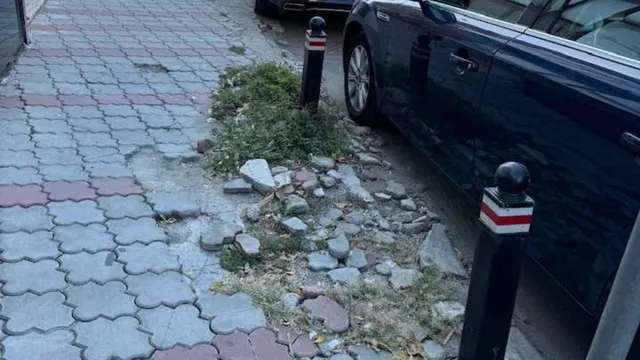 Un trotuar din pavaj colorat prezintă zone cu pietre de pavaj scoase și resturi de beton lângă un bolard și un autoturism albastru.