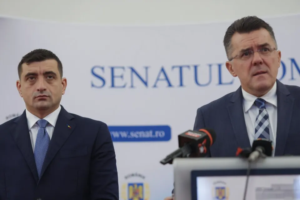 Un bărbat cu ochelari și cravată albastră stă în spatele unor microfoane în fața unui panou alb cu textul SENATULUI.