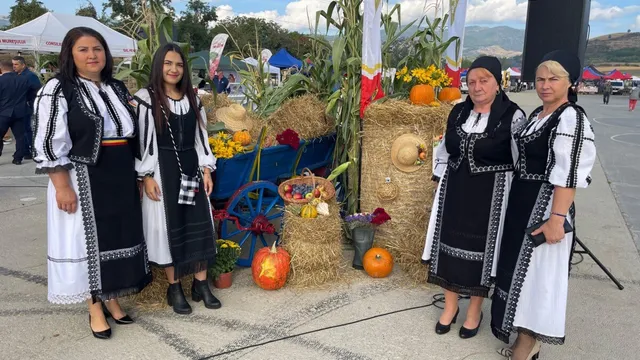 Patru femei poartă costume tradiționale albe cu elemente negre și stau în fața unor coșuri cu fructe, dovleci și stocuri de fân.