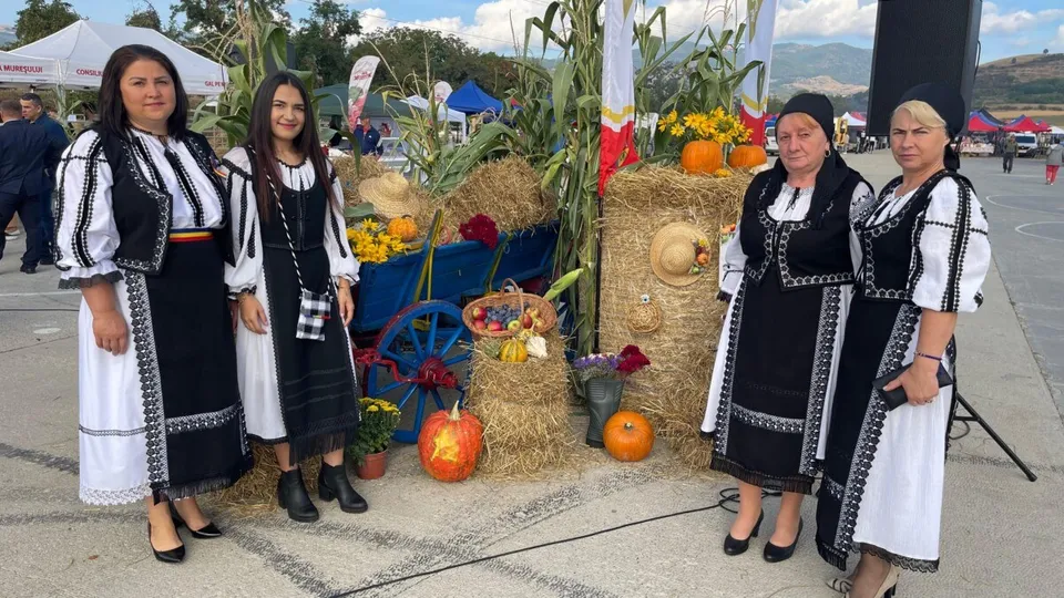 Patru femei poartă costume tradiționale albe cu elemente negre și stau în fața unor coșuri cu fructe, dovleci și stocuri de fân.